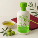 Bottle of Morellana olive oil with a green cap and label, surrounded by olives and a bowl of oil on a light background.