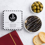 Can of agujas (needle-like pickled anchovies) with packaging and olives on a white surface