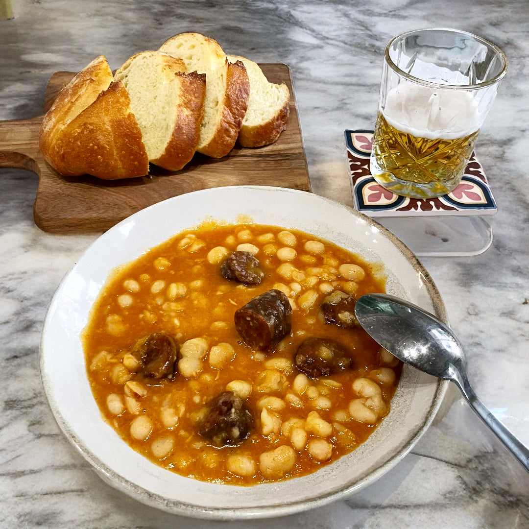 a plate of white beans with chorizo and some bread with a beer