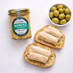 Jar of Bonito del Norte tuna, bread slices with tuna, and a bowl of green olives on a white background