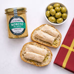 Jar of Bonito del Norte tuna, bread slices with tuna, and a bowl of green olives on a white background
