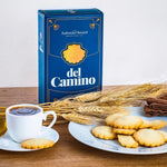 A box of 'Antonio Anaut del Camino' Spanish cookies with Conchas shape placed next to a plate of cookies, a cup of coffee, and some wheat ears on a wooden surface.