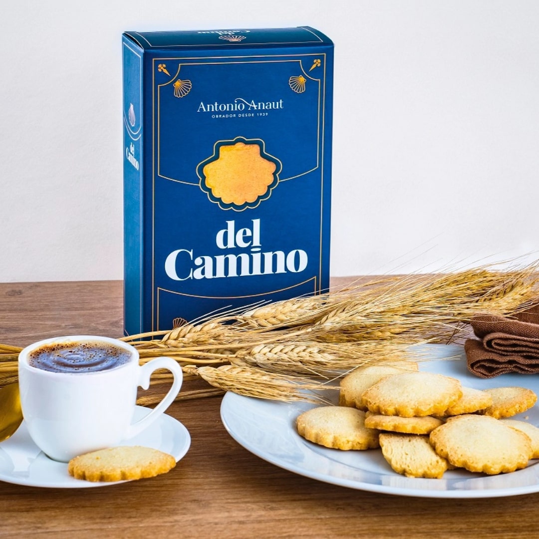A box of 'Antonio Anaut del Camino' Spanish cookies with Conchas shape placed next to a plate of cookies, a cup of coffee, and some wheat ears on a wooden surface.