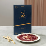 Cinco Jotas packaging with a plate of sliced iberico ham on a neutral background