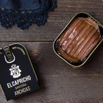 A tin can of El Capricho Cantabrian Anchovies in EVOO on a wooden surface, with the brand's logo visible on the lid.