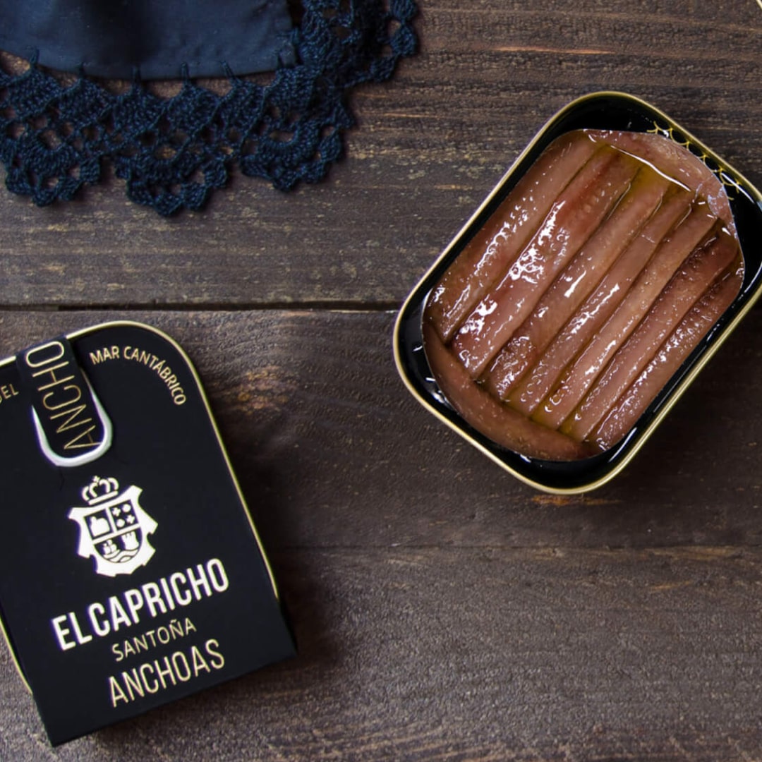 A tin can of El Capricho Cantabrian Anchovies in EVOO on a wooden surface, with the brand's logo visible on the lid.