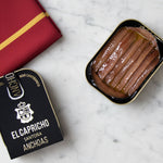 A tin can of El Capricho Cantabrian Anchovies in EVOO on a wooden surface, with the brand's logo visible on the lid.