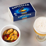 A can of Espinaler mussels in escabeche next to a bowl of mussels and a glass of beer on a grey surface.