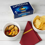 A can of Espinaler mussels in escabeche next to a bowl of mussels and a glass of beer on a grey surface.