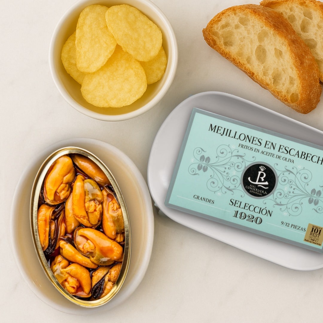 Canned mussels, potato chips, bread, and a can of 'Mejillones en Escabeche' on a white surface.