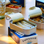 A tin can of Espinaler Razor Clams Navajas opened to display the large, meaty razor clams inside, placed on a wooden surface with other tins in the background.