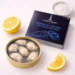 Oysters in a tin with a box labeled 'La Mar de Tazones' on a white surface with lemon slices and salt.
