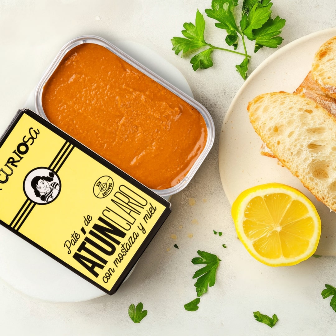 Can of bonito pâté La Curiosa with a yellow label, bread, lemon, and parsley on a white surface