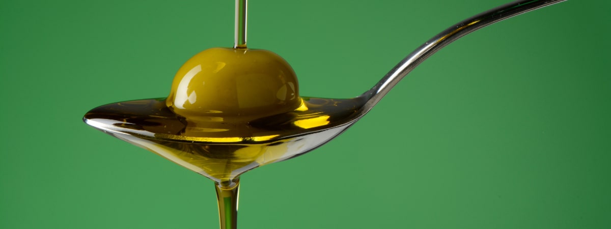 Olive oil being drizzled onto a green olive on a spoon against a green background