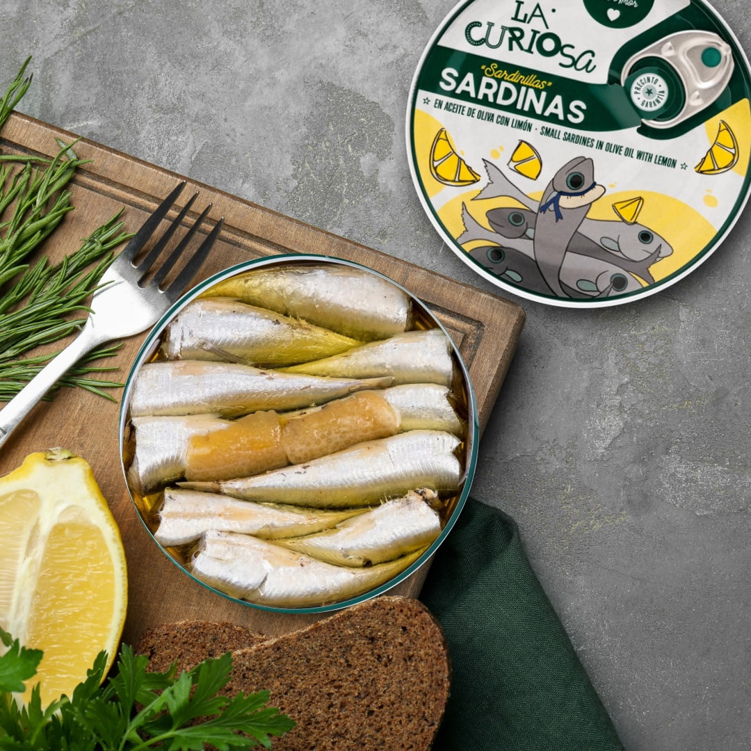 Tin of sardines on a wooden board with lemon, bread, and herbs, next to a 'La Curiosa' sardine can.