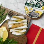 Tin of sardines on a wooden board with lemon, bread, and herbs, next to a 'La Curiosa' sardine can.