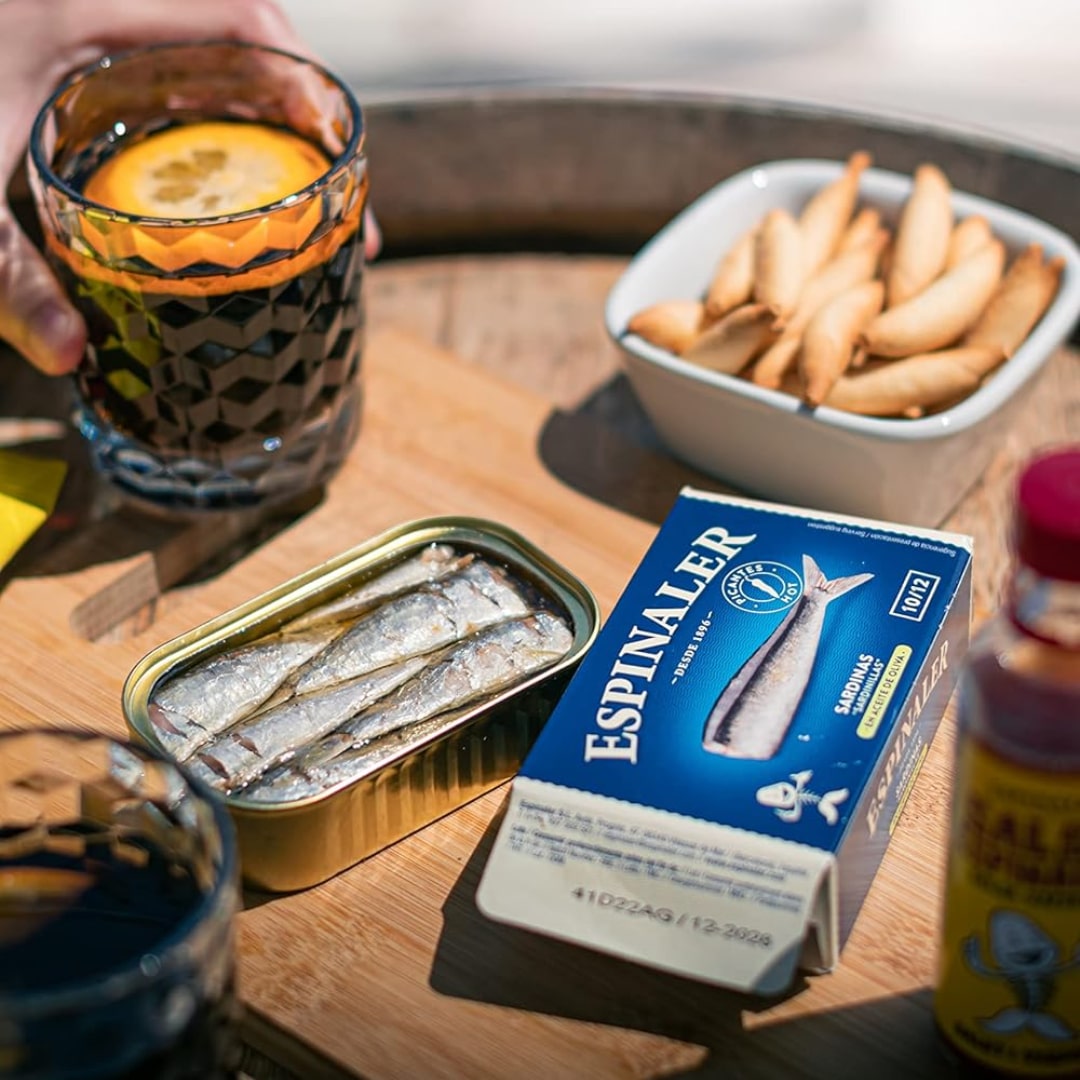A tin can of Espinaler small sardines in spicy olive oil, with a glass of lemon-infused drink and a bowl of toasted bread slices in the background, suggesting a gourmet seafood meal.