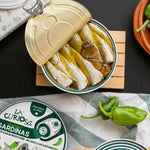 Open can of small sardines with padron peppers with a decorative label on a wooden coaster.