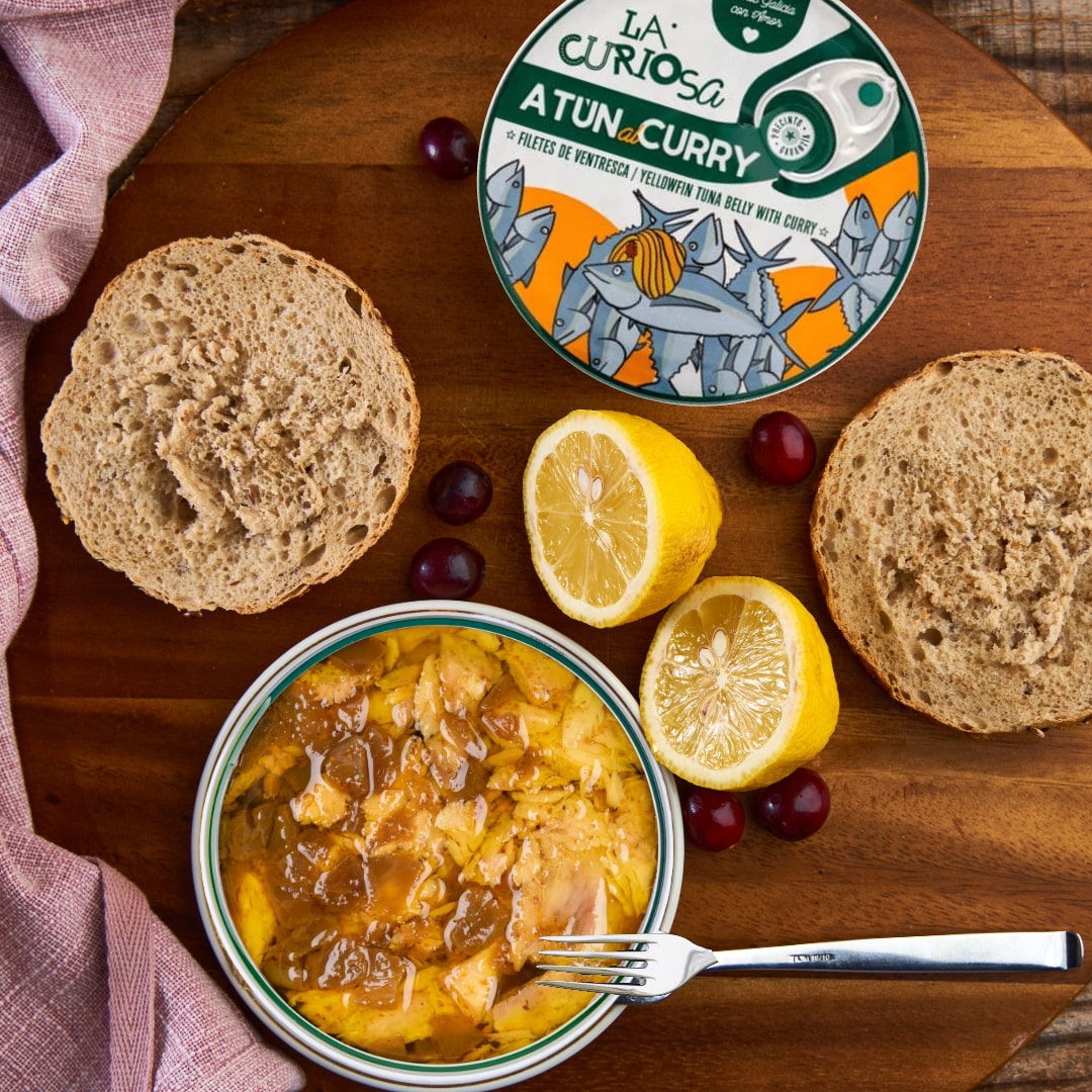 Bowl of tuna curry with bread, lemons, and grapes on a wooden board with La Curiosa Atun Curry packaging.