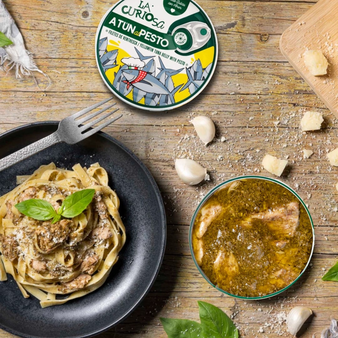 Pasta dish with pesto, a bowl of soup, and a container of La Curiosa Atun de Pesto on a wooden table.