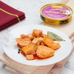 A plate of pickled white tuna, also known as Bonito Tuna in Escabeche, served on a white plate with a can of the product visible in the background. The tuna appears to be in a red pickled sauce, and there is a leaf of parsley garnish on the side.