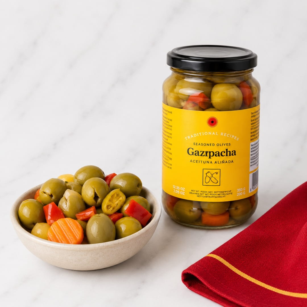 A jar of Gazpacha seasoned olives with red peppers and carrots stands next to a small bowl filled with sliced olives, topped with a black lid, on a white marble surface with a red napkin.