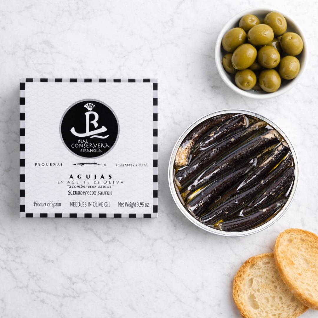 Can of agujas (needle-like pickled anchovies) with packaging and olives on a white surface