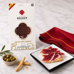 Beher jamón packaging with sliced ham on a plate, olives, and bread sticks on a marble surface.