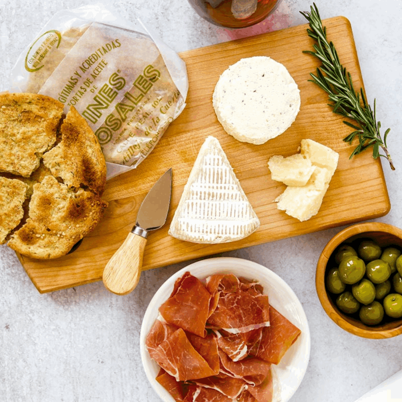 table with ines rosales tortas, a piece of cheese, a plate with serrano ham and a bowl with green olives