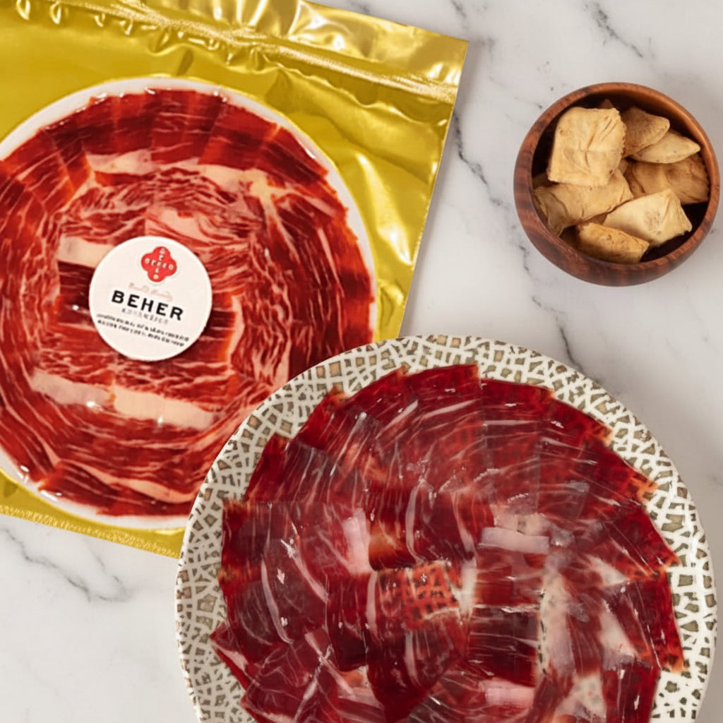 Two packages of sliced jamon iberico with a Beher label, one open showing the slices, on a light surface with a bowl of small items.