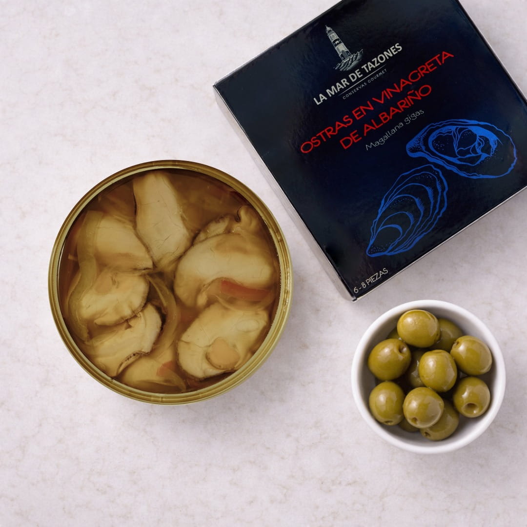 Oysters in a can with a box labeled 'La Mar de Tazones' and a bowl of green olives on a light surface.
