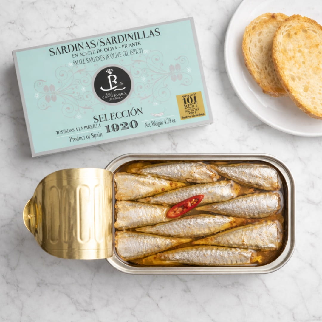 Open can of sardines with a lid on a marble surface, next to a box labeled 'Sardinas Sardinillas'.