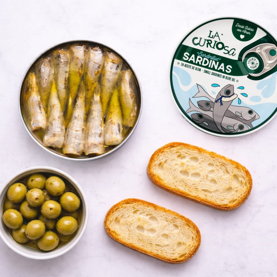 Canned sardines, green olives, and bread slices on a white surface with 'La Curiosa' packaging.