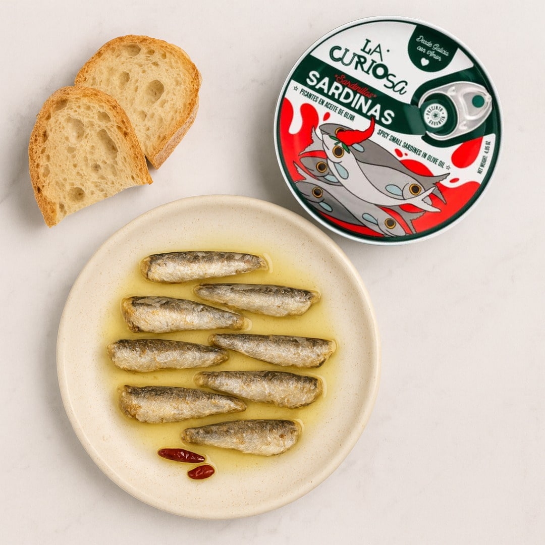Sardines on a plate with bread and a can of sardines labeled 'La Curiosa' on a light background.